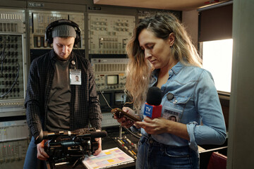 Young reporter with microphone using smartphone while standing by workplace and male operator...