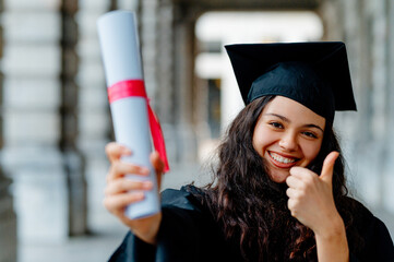 Young latin woman wearing a graduation gown, proudly displaying her diploma and giving a thumbs up in celebration of her achievement