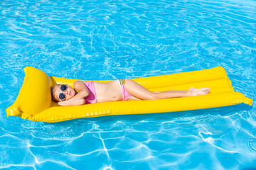 Cute little girl lying on inflatable mattress in swimming pool with blue water on warm summer day...