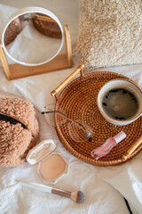 Cup of hot coffee on wicker rattan tray, boucle cosmetic bag, cosmetics on white bed linen.