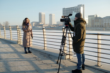 Young man standing in front of professional video camera on tripod while shooting female journalist...