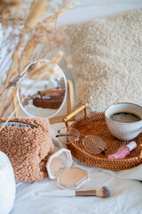 Cup of hot coffee on wicker rattan tray, boucle cosmetic bag, cosmetics on white bed linen.