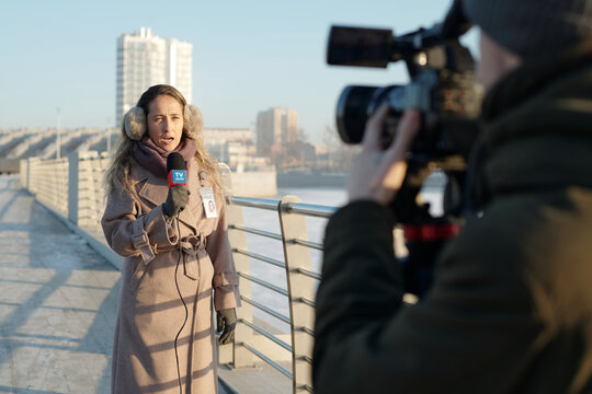 Young confident woman in warm beige coat and earmuffs speaking in microphone while reporting about latest events