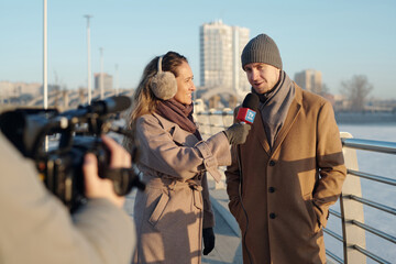Fototapeta premium Young man and woman in beige coats standing in front of video camera shooting outdoor interview by riverside