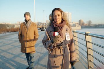 Young female reporter in winterwear looking at camera and speaking in microphone against man...