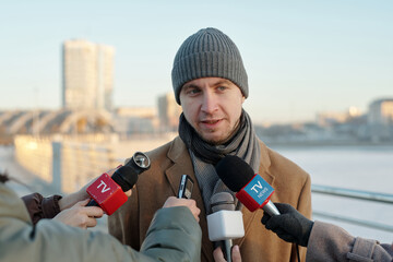 Young confident man in coat and beanie hat speaking in microphones in hands of reporters of several TV channels outdoors