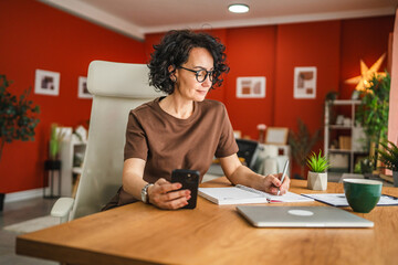 woman work from home and take notes in notebook while use mobile phone