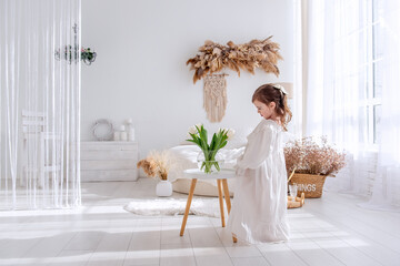 Little girl in a white dress, nightgown. Toddler collects bouquet of fresh, delicate white tulips in a round glass vase on a wooden table. Light, trendy interior in boho style. Copy space. Spring time