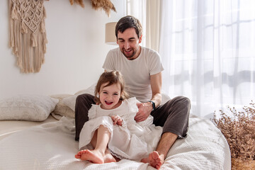 Happy father tickling his cheerful daughter while sitting together on a comfortable bed in a cozy,...