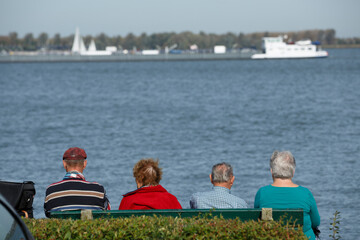 Elderly friends enjoying a sunny afternoon by the river while watching boats sail by in a peaceful park setting