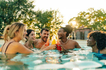 Friends toasting with colorful drinks in swimming pool at sunset