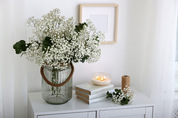 Bouquet of beautiful gypsophila flowers and eucalyptus branches in vase near decorative elements on chest of drawers indoors