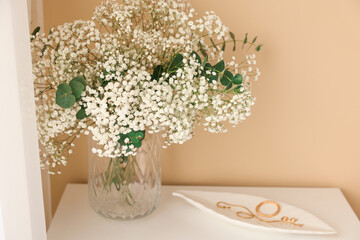 Bouquet of beautiful gypsophila flowers and eucalyptus branches in vase on chest of drawers near beige wall