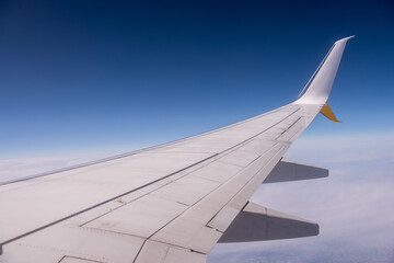 The wing of an airplane slices through the clear blue sky, offering a breathtaking view of the world from above, with a thin layer of clouds far below