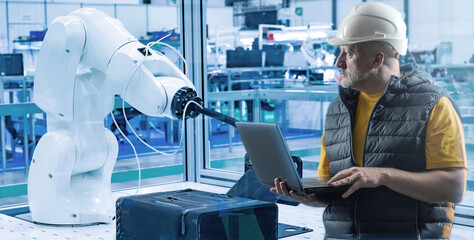 Man industrial automation engineer. Arm manipulator behind worker. Man controls industrial robot via laptop. Arm manipulator in automated factory. Engineer controls robotic production