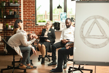Group of alcoholics gather in aa meeting room with symbol displayed on white board. Retired...