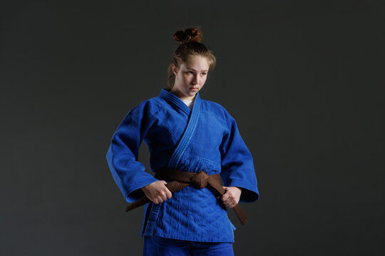 Determined young female martial artist in a blue gi tying her brown belt, focused expression, isolated on dark background.