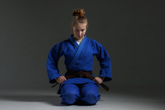 Determined young female martial artist in a blue gi tying her brown belt, focused expression, isolated on dark background.