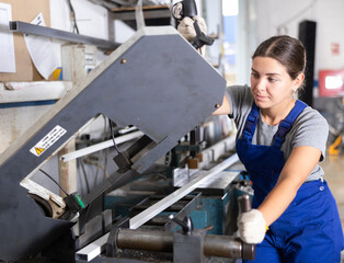 Confident young woman in blue overalls and gloves cutting PVC profile on industrial machine in...
