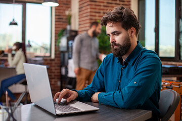 Fototapeta premium Male employee typing in business plans on his laptop at desk in brick wall workspace. Caucasian office worker seated at table, researching financial and marketing strategies for startup company.