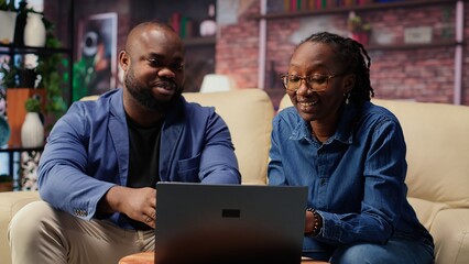 Black couple in living room discusses creative ideas for an online business, using modern...
