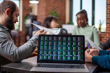 Colleagues discuss in brick wall office, reviewing documents as the laptop on table displays financial statistics. Digital device on desk showing market analysis data in modern workspace.