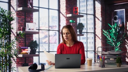 Casual focused woman sitting at her desk and reading research papers, multitasking with laptop at...