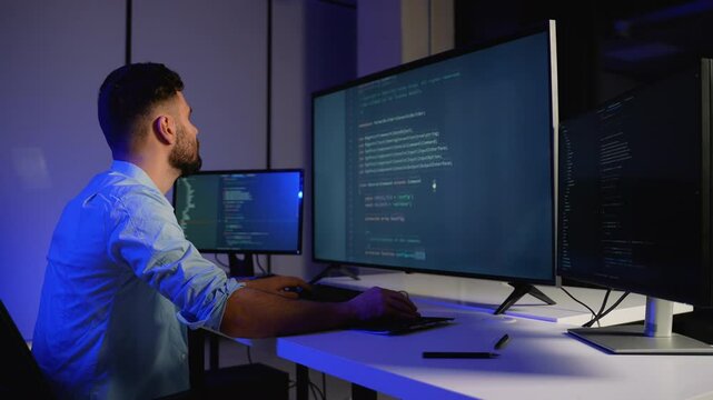 Programmer writing code on multiple screens in a dark office