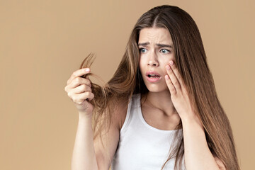 Young girl with long hair looking at her split ends in panic, beige studio background