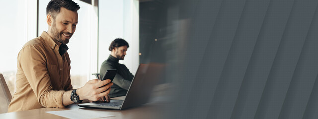 Happy man company employee checking smartphone, using laptop, working with male colleague at modern office, free space. Professional occupation, business success concept