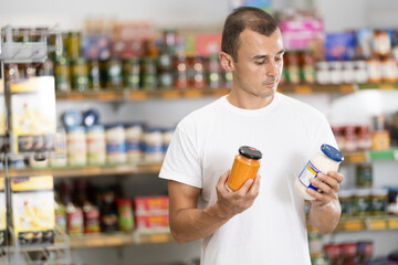 Guy in store chooses interested in sauces, examines information on pack, gets acquainted with assortment. Buyer compares different product lines, looks at packages of different sauces