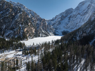 Aerial view of Braies Lake in winter, surrounded by snow covered forests and rocky peaks. A lodge sits near the frozen lake under a clear blue sky.