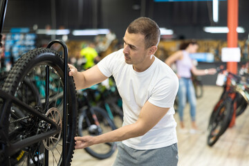 Young male customer choosing bike and checking tire on bicycle wheel in modern sport store