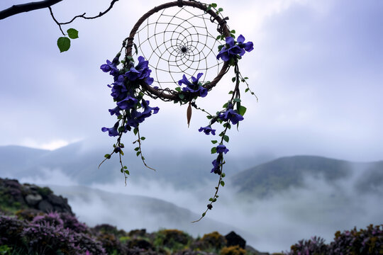 Dreamcatcher with purple flowers hanging on a branch. Misty mountains and cloudy sky create a peaceful, nature-filled scenic view.