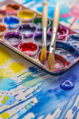 Obraz premium A vibrant and dynamic scene of a watercolor painting in progress. An open palette filled with colorful pigments sits atop a textured watercolor paper covered in expressive splashes and brush strokes.