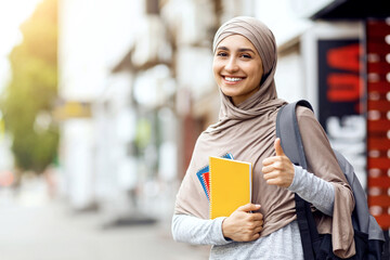 Pretty arab girl with notepads and books showing thumb up, standing on the street, copy space