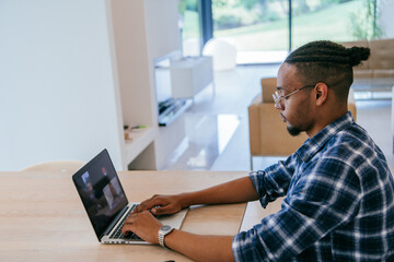 African American influencer in glasses sitting at a table in a modern living room, using a laptop for business video chat, conversation with friends and entertainment.