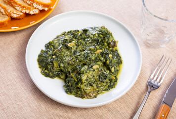 On table there is plate with stewed side dish, spinach in Catalan. Dish is steamed with addition of pine nuts and raisins. It is served with meat and fish dishes