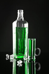 Absinthe in shot glass, bottle and ice cubes on black mirror surface