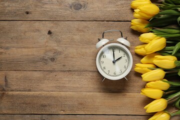 Spring time. Alarm clock and beautiful tulips on wooden table, flat lay. Space for text