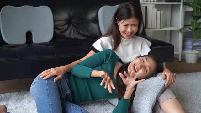 Two cheerful Asian women. Likely friends or sisters. Are enjoying a lighthearted moment. Engaging in playful tickling on the floor of a cozy living room. Creating a scene of genuine joy and amusement