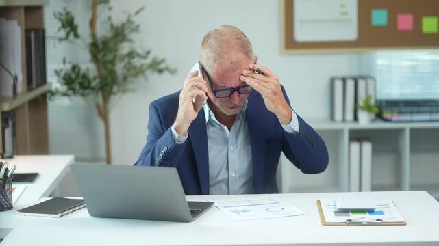 A man in a suit is talking on his cell phone while wearing glasses. He is sitting at a desk with a laptop and a stack of papers. Concept of busyness and focus