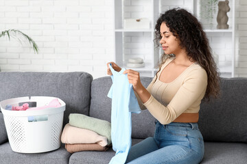 Beautiful young happy African-American woman with laundry basket folding clean clothes at home
