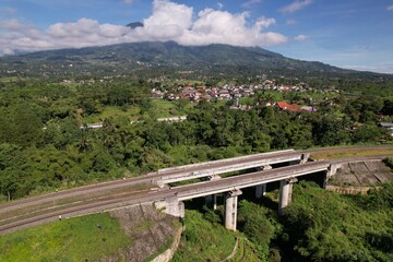Naklejka premium Aerial view of a railway bridge over a lush valley, with a village at the foot of a cloud-covered Mount Salak in Bogor. A perfect blend of nature and infrastructure in Indonesia’s highlands