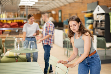 Girl buyer before buying furniture inspects chair goods for defects and flaws. Customer in garden furniture store inspects countertop and imagine product in patio