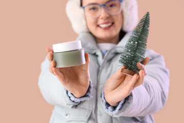 Young woman in winter clothes with jar of cream and toy Christmas tree on beige background, closeup