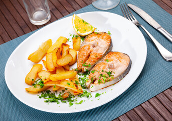 Portion of just cooked grilled salmon steak garnished with fried potato, served on plate with parsley and piece of lemon.