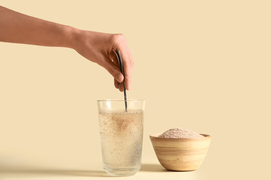 Female hand with spoon and psyllium husk in glass of water on beige background