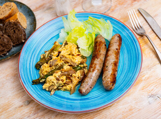 Scrambled eggs with mushrooms, chicken fillet and asparagus are served on plate. Frankfurter sausages and slices of fresh soft bread are added to fried eggs, and china cabbage leaf
