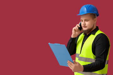Male engineer with clipboard talking by mobile phone on dark red background